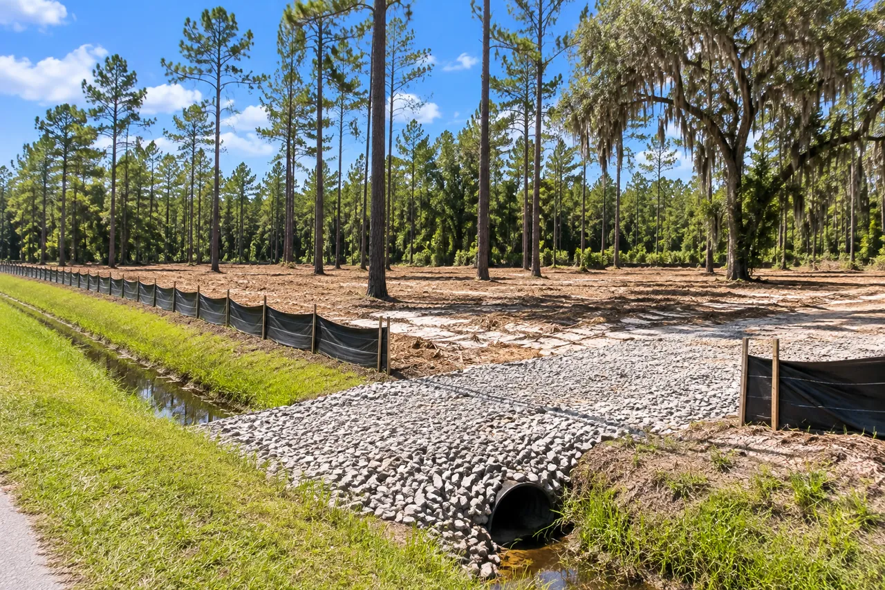 Black silt fence along a cleared sandy lot beside a grassy roadside ditch, with a stone construction entrance